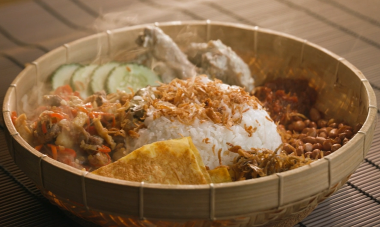 A wooden bowl with rice, fried chicken, anchovies, peanuts, sliced cucumber, egg, and sambal chili mixture.